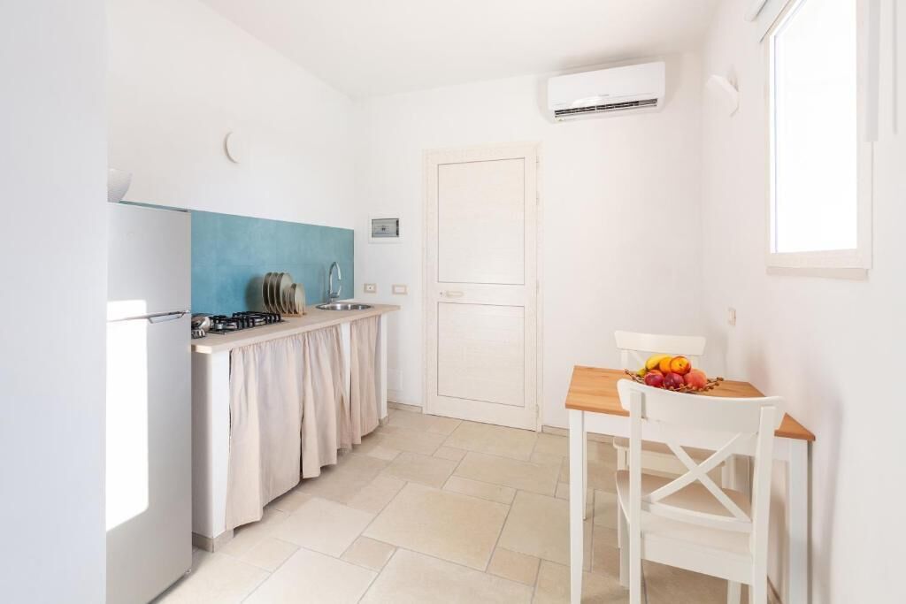 Una cocina compacta con estufa a gas, un fregadero y un salpicadero azul. Hay una zona de comedor con una mesa y sillas, y una puerta que da a otra habitación.
