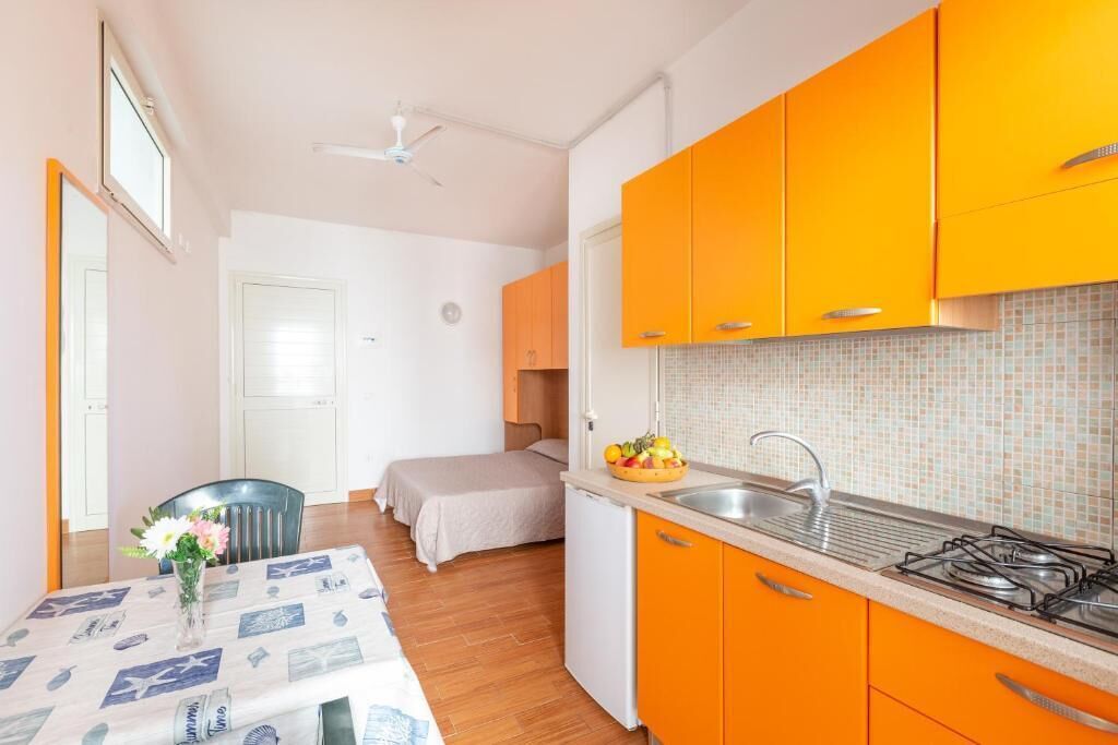 A compact kitchen with orange cabinets, a small sink, and a stove. There is a dining area with a table and chairs, and a bed in the background.