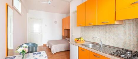 A compact kitchen with orange cabinets, a small sink, and a stove. There is a dining area with a table and chairs, and a bed in the background.