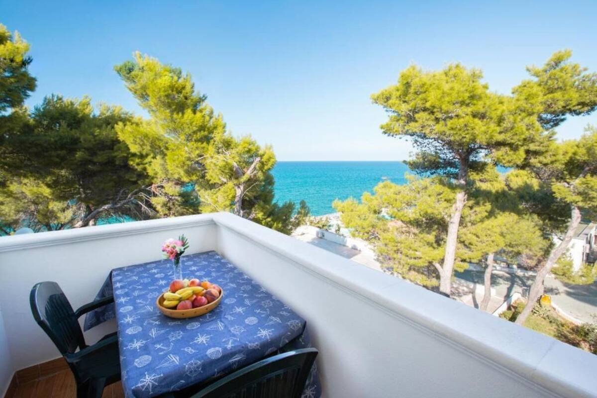Un balcon avec une table dressée pour deux personnes, offrant une vue sur la côte, avec des arbres et la mer.