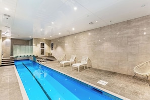 An indoor swimming pool with clear water, surrounded by beige tiled walls and white chairs.
