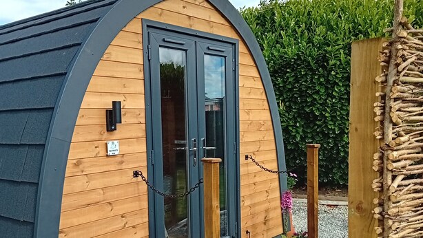 A small, wooden, semi-circular structure with a dark roof and a wooden deck leading to a glass door.
