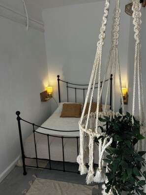 Een bed met metalen frame, twee bedlampen en een hangstoel van macramé.