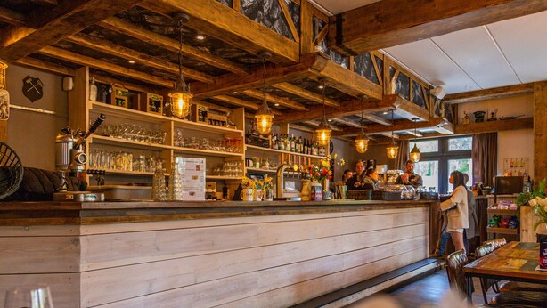A rustic bar area with wooden beams, shelves stocked with bottles and glasses, and a counter where a bartender is serving. - Tent Lodge With Bathroom 16 km From Maastricht (Zutendaal)