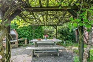 Ein rustikaler Außenbereich mit Holztisch und Bänken unter einer Pergola, umgeben von üppigem Grün.