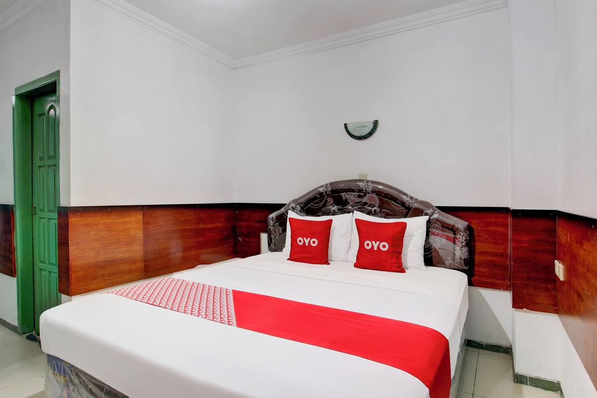 Quarto de hotel com cama, porta verde, paredes brancas e um relógio na parede.