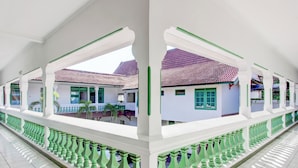 Un couloir couvert, orné de colonnes blanches et de balustrades vertes, mène à un bâtiment dont le toit est recouvert de tuiles rouges.