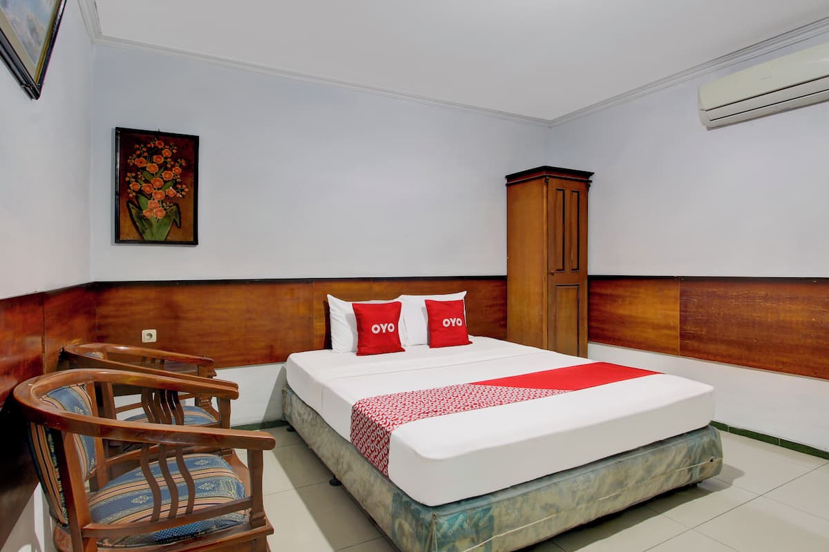Quarto de hotel com cama, cabeceira de madeira, guarda-roupa de madeira, uma cadeira e um quadro emoldurado na parede.