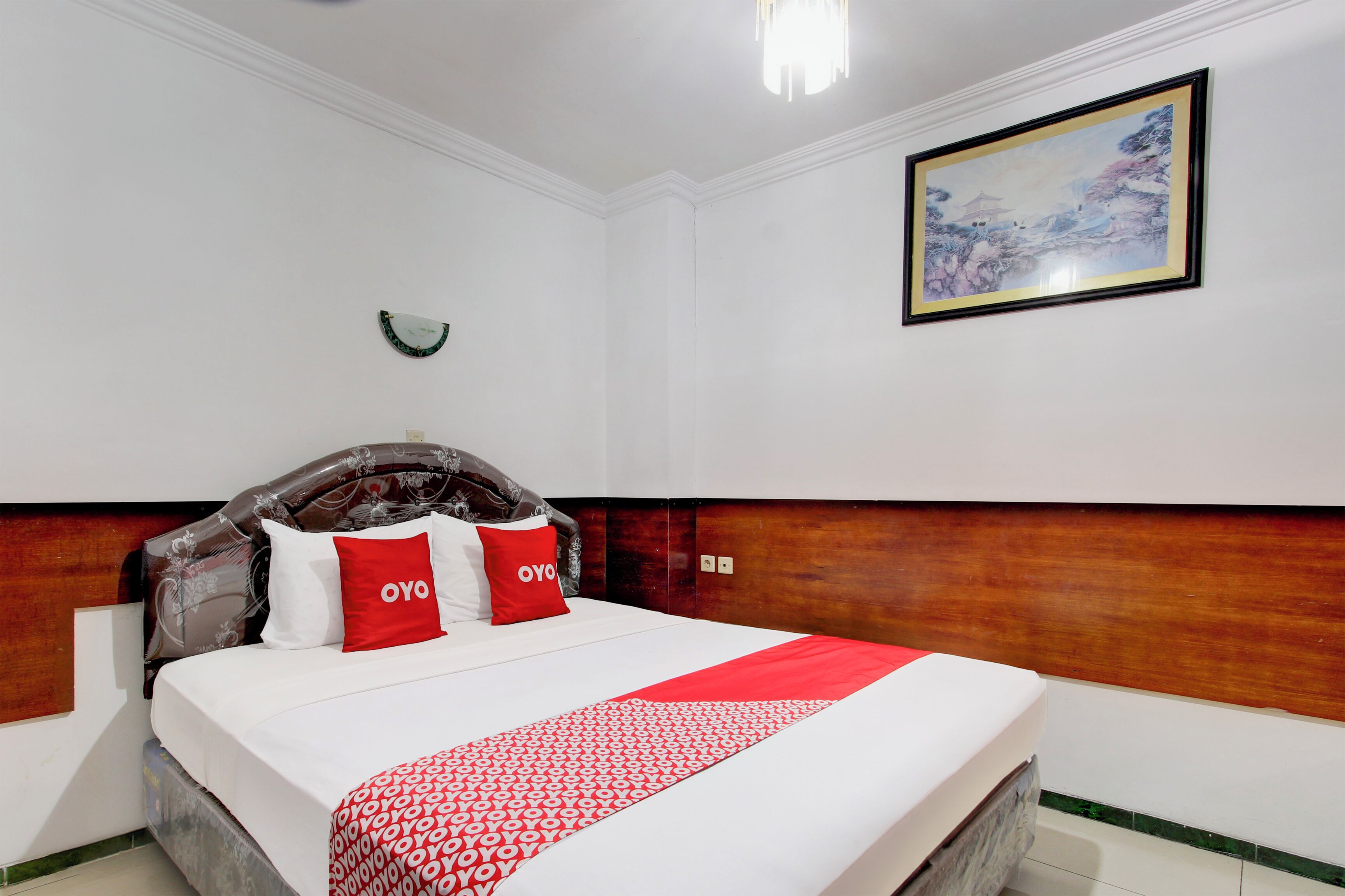 A hotel room with a bed, a wooden headboard, a red and white bedspread, and a framed picture on the wall.