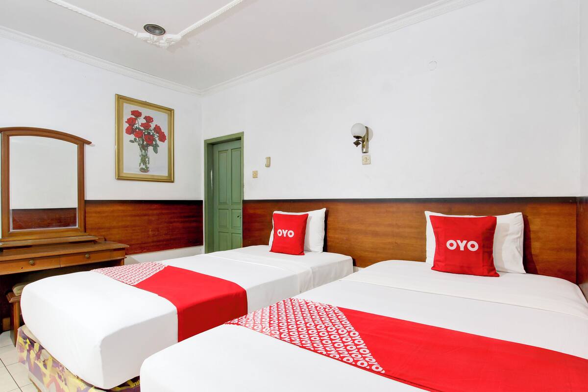 Quarto de hotel com duas camas, cada uma com colcha e almofadas vermelhas e brancas. Há uma cômoda de madeira, uma porta verde e um quadro na parede com flores vermelhas.