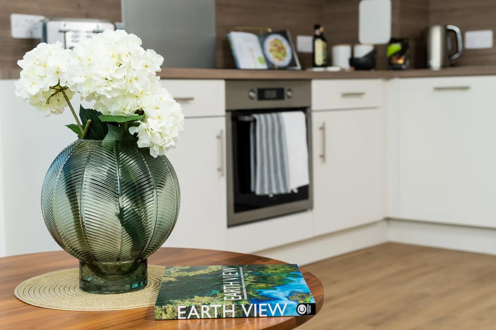 Eine moderne Küche mit weißen Schränken, einem Edelstahlbackofen und einer Vase mit weißen Blumen auf einem Holztisch.