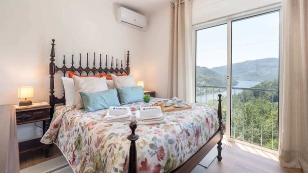 A bedroom with a large bed, a nightstand, a bedside lamp, a window with a view of the outdoors, and a balcony.