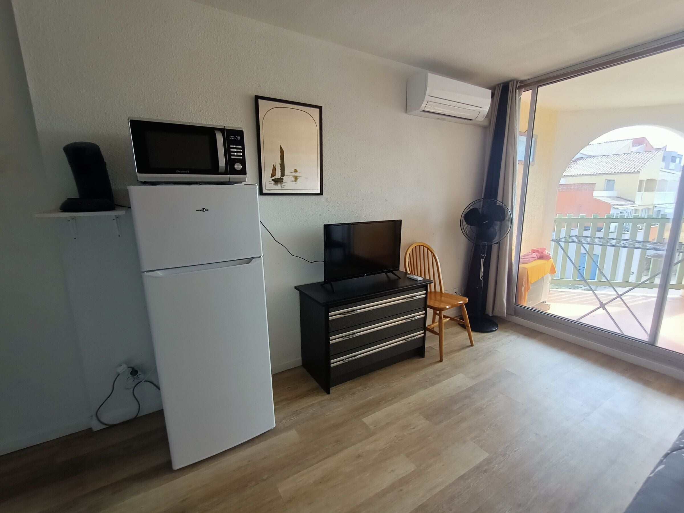 A compact living space with a refrigerator, microwave, and a flat-screen TV on a dresser. A chair is positioned near a window with a view of a balcony.