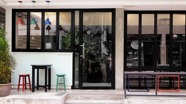 A modern storefront with large glass windows, a potted plant, and a small outdoor seating area with stools.