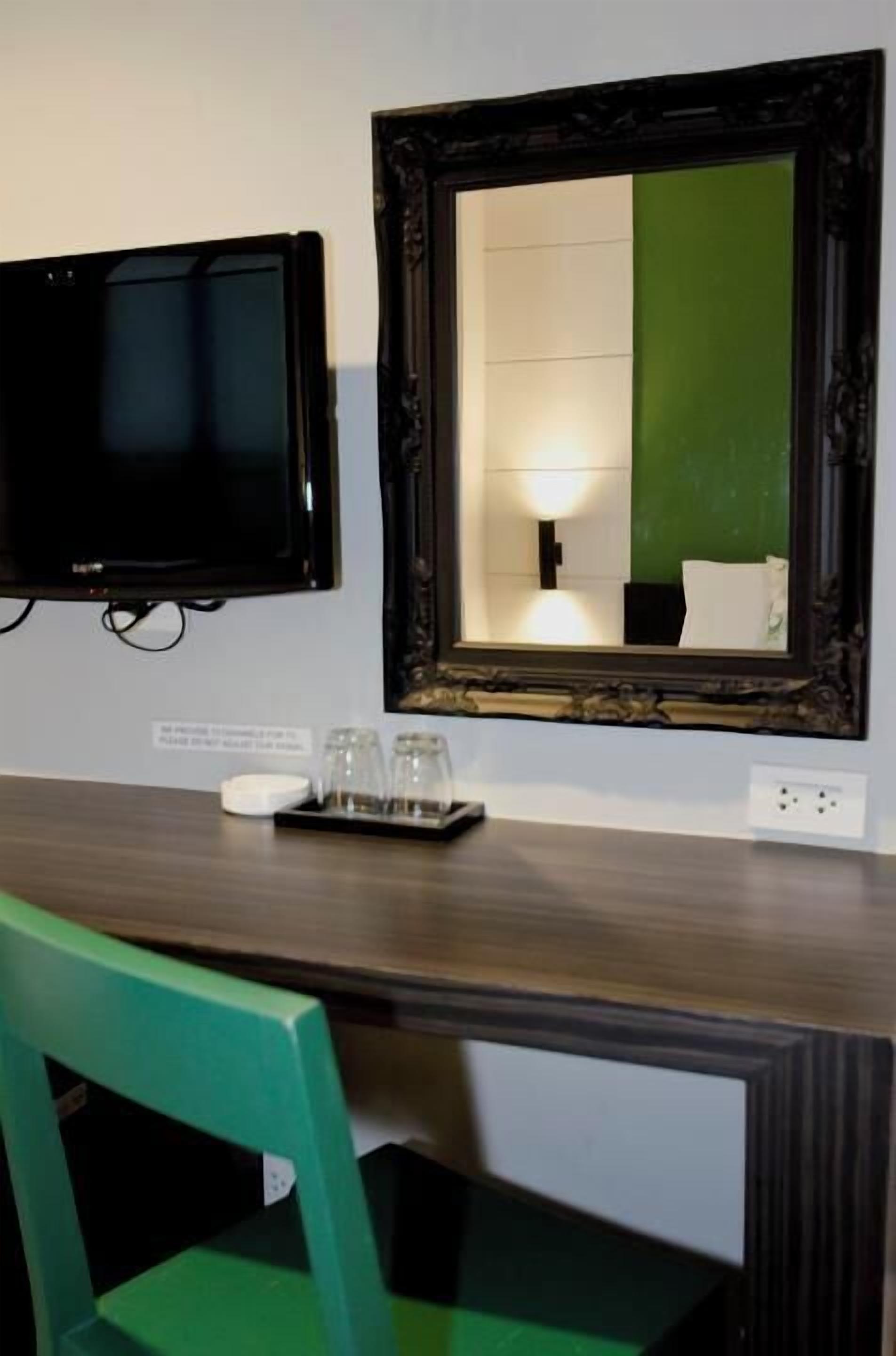 Une chambre d’hôtel avec un téléviseur fixé au mur, un miroir doté d’un cadre décoratif, un bureau en bois sur lequel se trouve un plateau avec des verres, et une chaise verte.