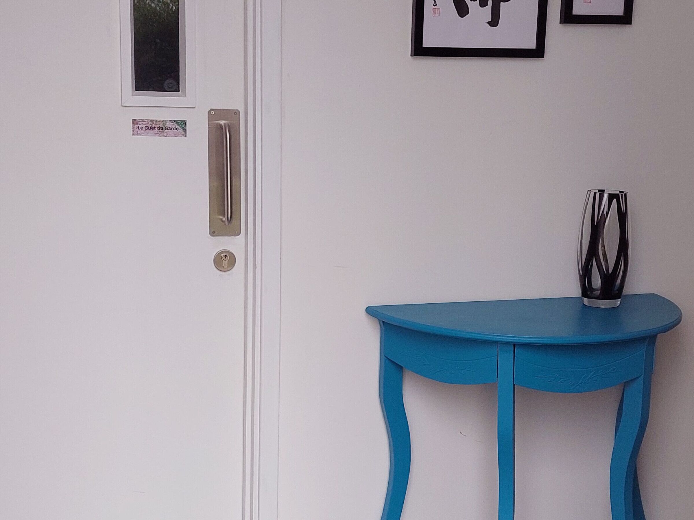 Une porte blanche avec une poignée argentée et un trou de serrure, un petit tableau encadré et une table bleue sur laquelle se trouve un vase.