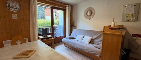 A room with a bed, a table, a TV, a wooden desk, a window with curtains, and a wall clock.
