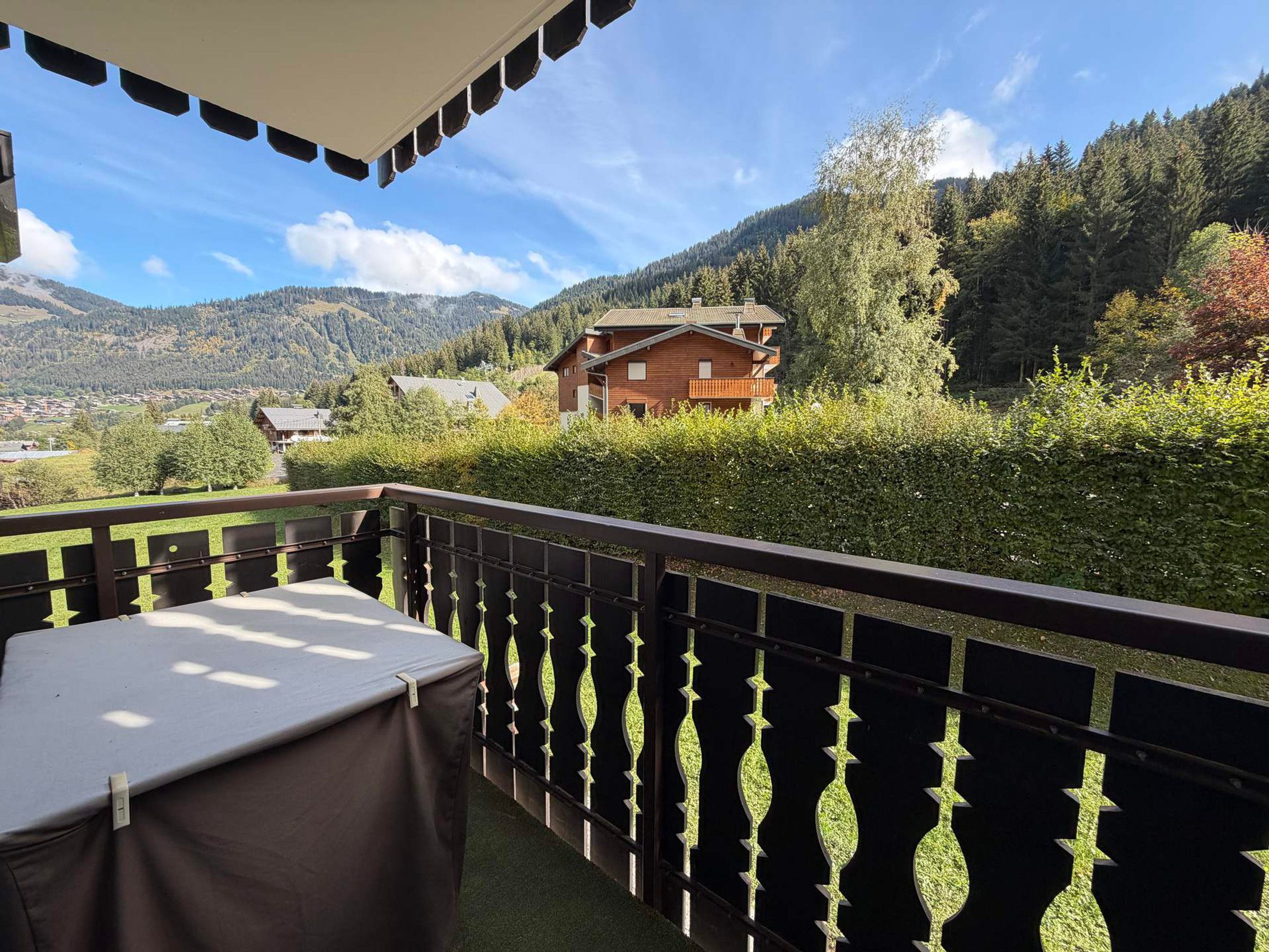 Un balcon avec une table et des chaises, donnant sur un paysage montagneux où l’on aperçoit, au loin, un bâtiment en bois.