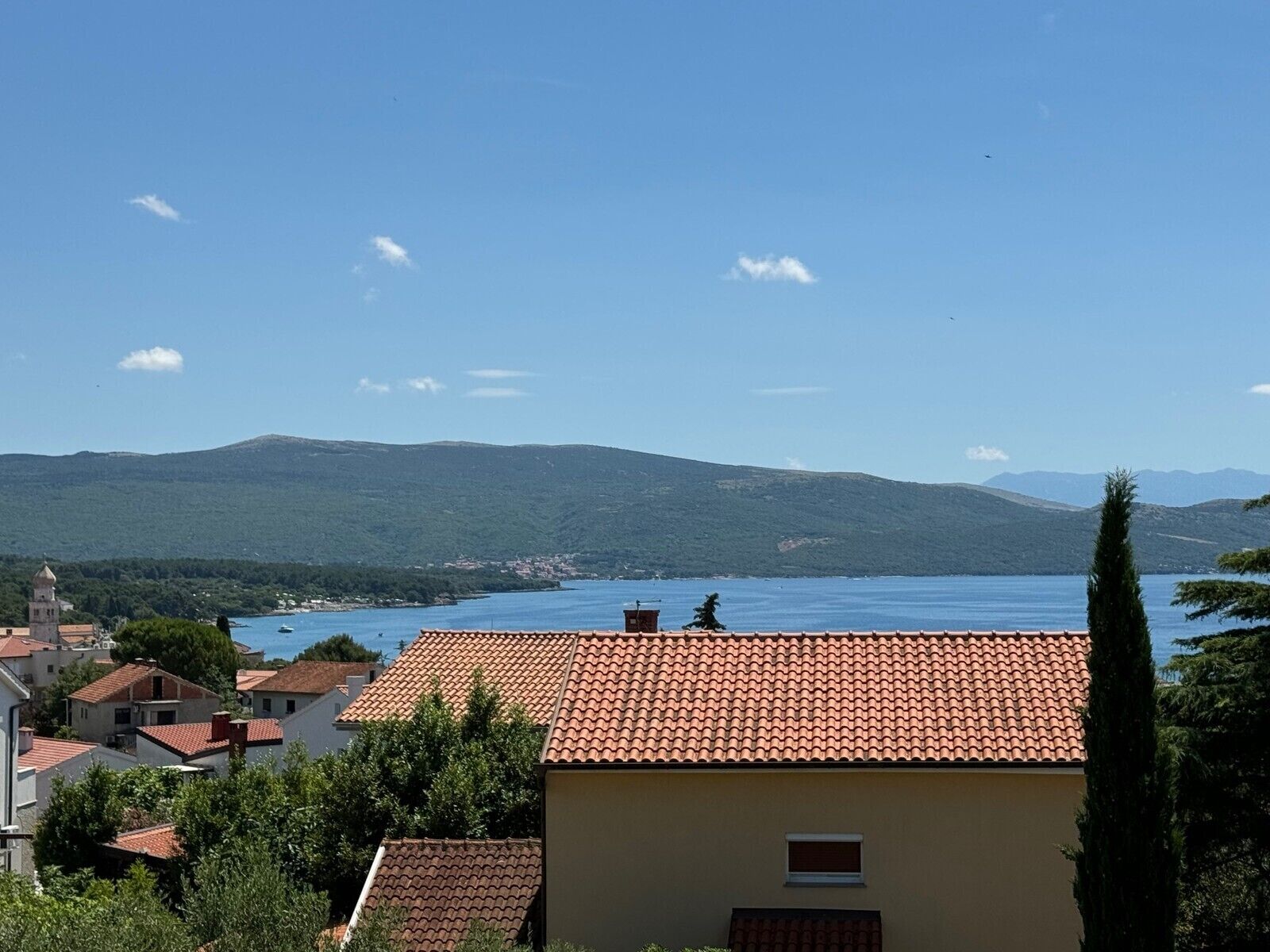 A coastal town with terracotta-roofed buildings, a clear blue sky, and a view of the sea and distant mountains.
