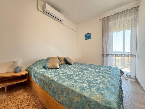 Un dormitorio con cama, mesita de noche, lámpara, aire acondicionado, ventana con cortinas y un pequeño cuadro en la pared.