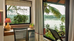 A room with a view of a body of water, a desk with a lamp, a chair, and a small refrigerator. - Vedic Village Retreat (Kodungallur)