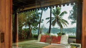A bedroom with a view of a lake, a bed with pillows, a small table with a basket, and a window with a view of palm trees. - Vedic Village Retreat (Kodungallur)