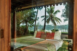 Un dormitorio con vista a un lago, una cama con almohadas, una mesita con una cesta y una ventana con vista a palmeras.