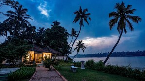 A serene lakeside setting with a small building, palm trees, and a pathway leading to the water. - Vedic Village Retreat (Kodungallur)