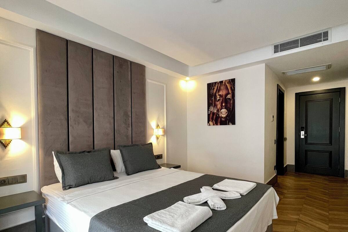 A modern hotel room with a large bed, grey bedding, a headboard, bedside lamps, and a painting on the wall.