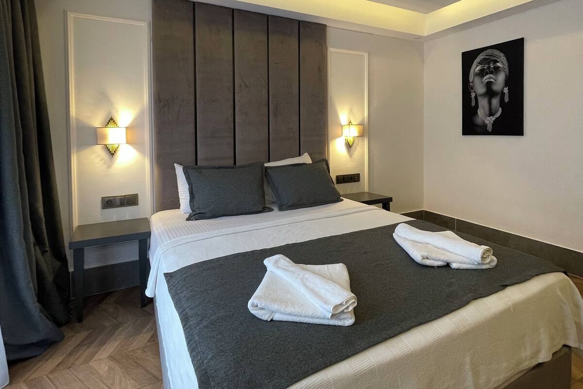 A modern hotel room with a large bed, two bedside tables with lamps, a headboard, and a wall-mounted artwork.