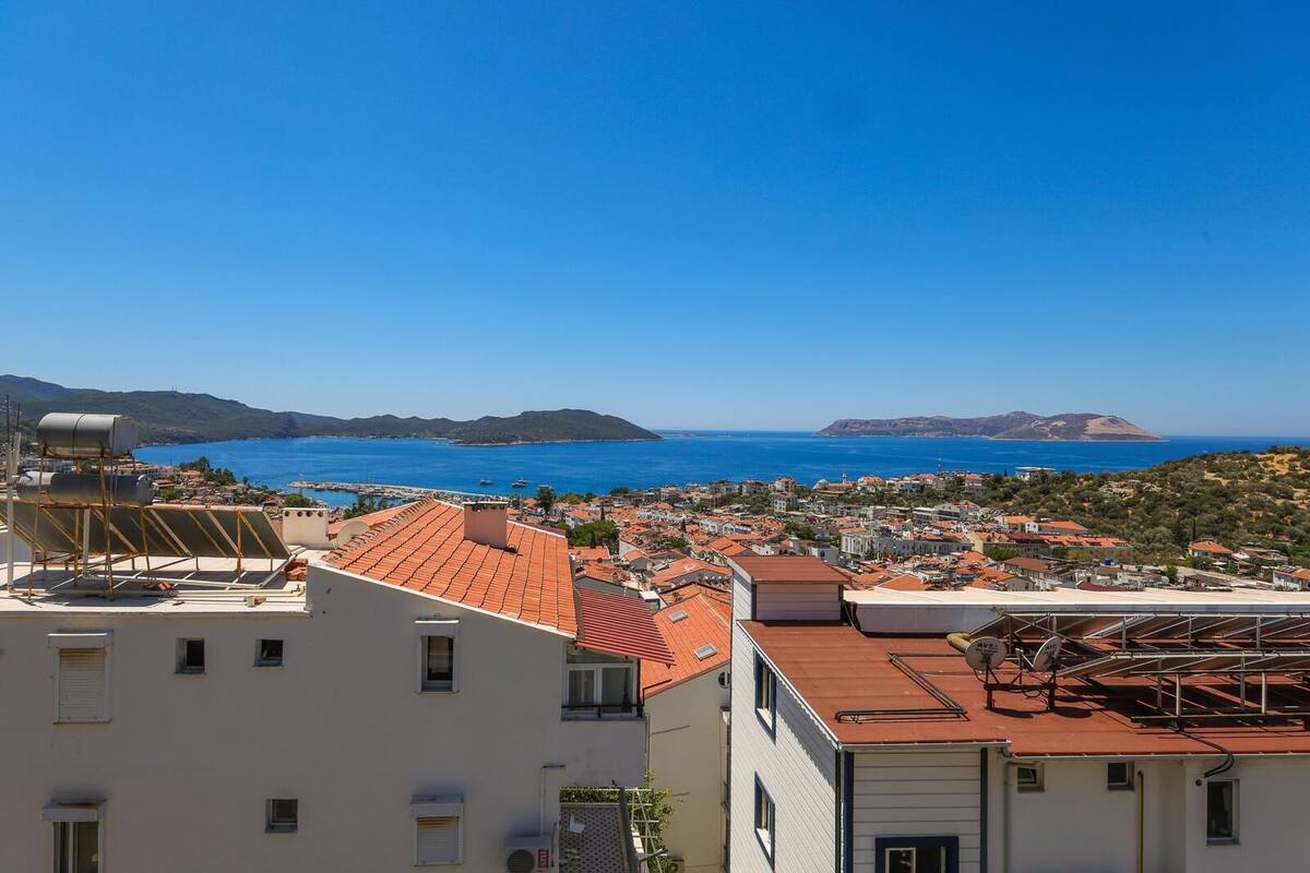 A coastal town with terracotta-roofed buildings, a clear blue sea, and distant hills.