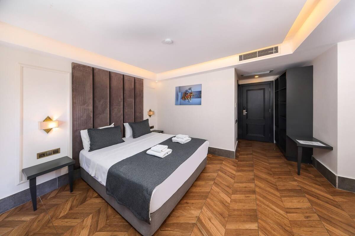 A modern hotel room with a large bed, wooden flooring, and a dark headboard.