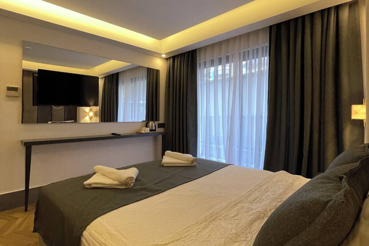 A modern hotel room with a large bed, a flat-screen TV mounted on the wall, a desk with a kettle, and a window with dark curtains.