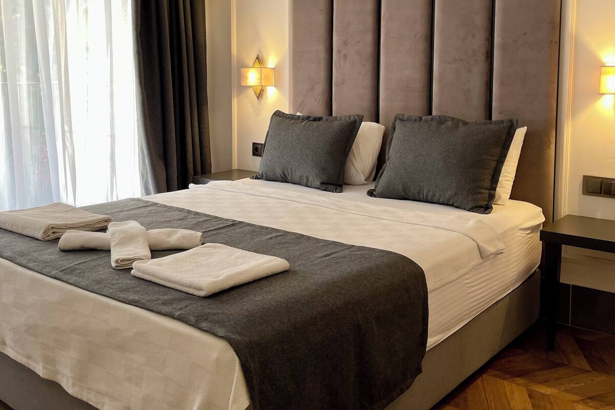 A modern hotel room with a large bed, a wooden herringbone floor, and a view of the outdoors through sheer curtains.
