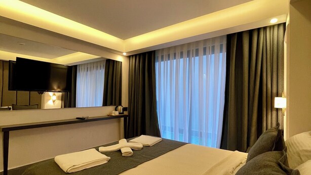 A hotel room with a bed, a flat-screen TV mounted on the wall, a bedside table with a lamp, and a window with curtains. - PİER HOUSE KAŞ (Kas)