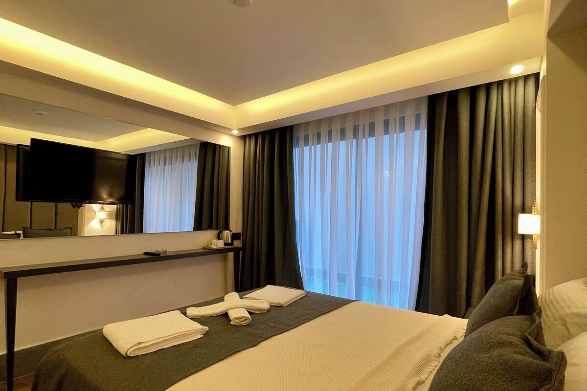 A hotel room with a bed, a flat-screen TV mounted on the wall, a bedside table with a lamp, and a window with curtains.