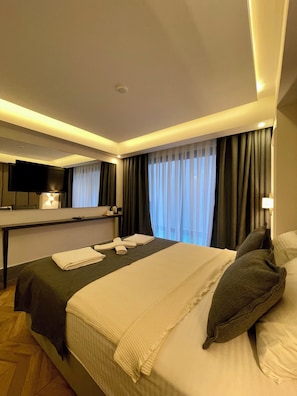 A hotel room with a bed, a flat-screen TV mounted on the wall, a bedside table with a lamp, and a window with curtains. - PİER HOUSE KAŞ (Kas)