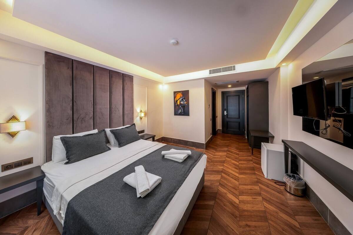 A modern hotel room with a large bed, bedside tables, a flat-screen TV, and a hallway leading to a door.