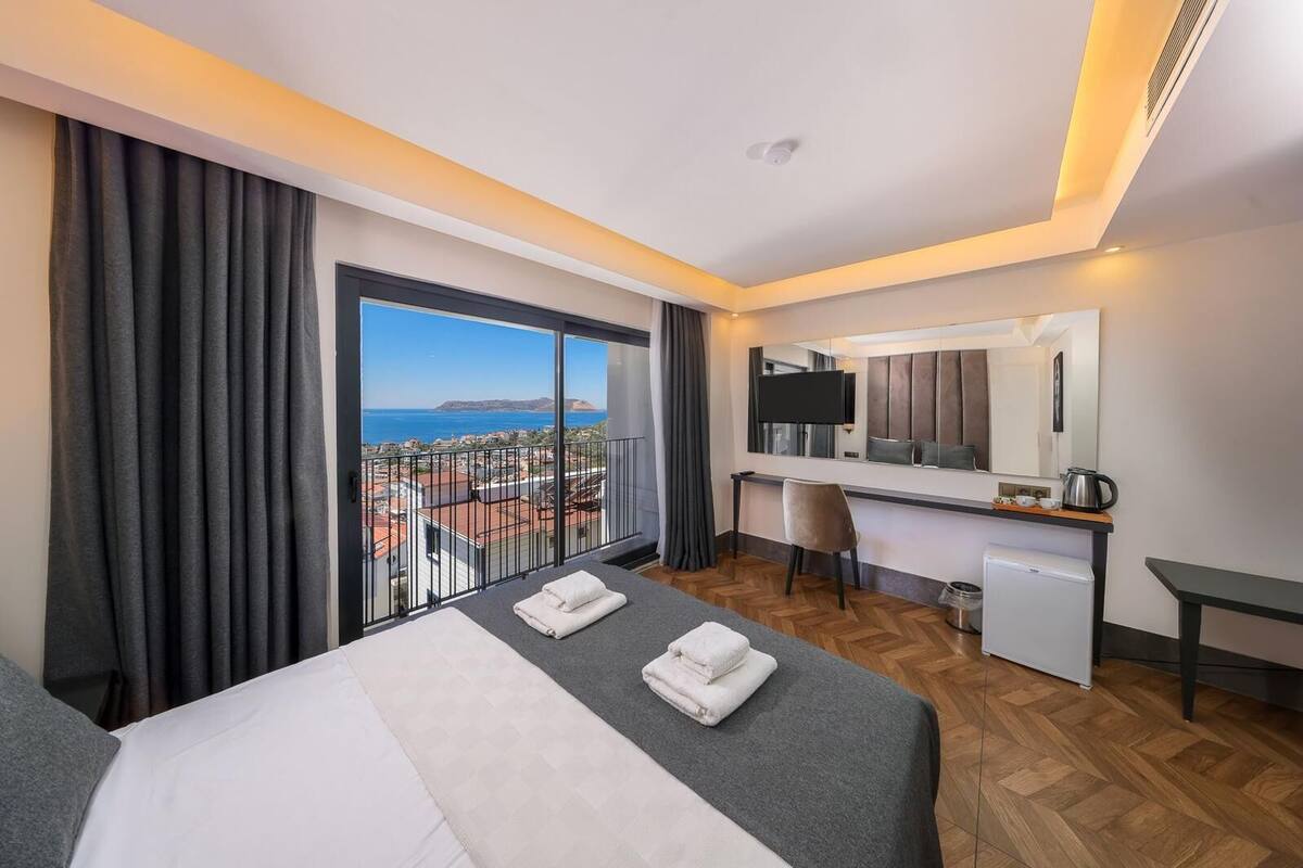 A hotel room with a bed, a desk, a chair, a small kitchenette, and a large window with a view of the city and sea.