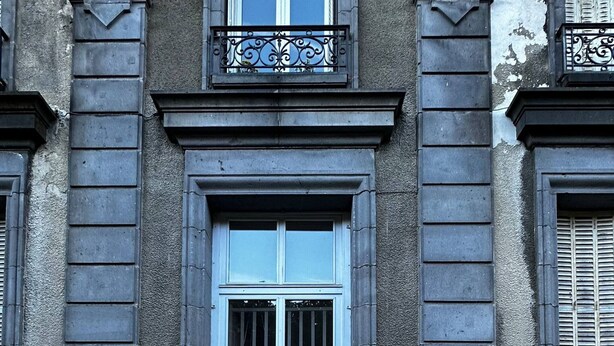 A building facade with a central entrance, a balcony with ornate metal railings, and a number '51' sign on the wall.