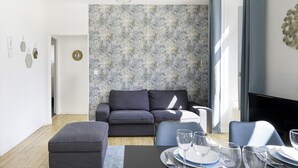 A modern living room with a gray sofa, a dining table set with plates and glasses, and a large window with curtains.