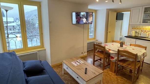A living room with a sofa, a coffee table, and a TV mounted on the wall. There is a dining area with a table and chairs.