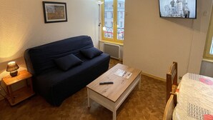 A furnished living room with a sofa, a coffee table, a TV mounted on the wall, and a window with a view of buildings.