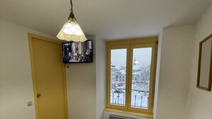 A bedroom with a bed, a television mounted on the wall, a window with a view of a snowy cityscape, and a small stool.