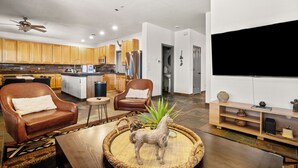 A modern kitchen with wooden cabinets, a central island, and stainless steel appliances. There is a dining area with a brown leather sofa and a coffee table. A flat-screen TV is mounted on the wall, and there is a small side table with a plant. - Serene desert escape w/ stunning mountain views & heated pool/spa- pet friendly! (Tucson)