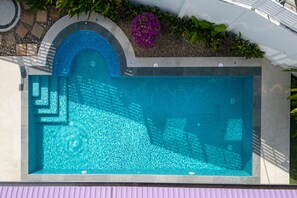 Una piscina rettangolare con una caratteristica di acqua cristallina e blu, circondata da un bordo in pietra e una piccola area giardino con fiori viola e piante verdi.