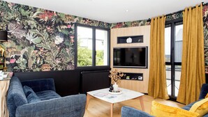 A living room with a large TV, a sofa, a coffee table, and a vibrant floral wallpaper. - La Maison des Dunes - Plage à Pied (Cléder)