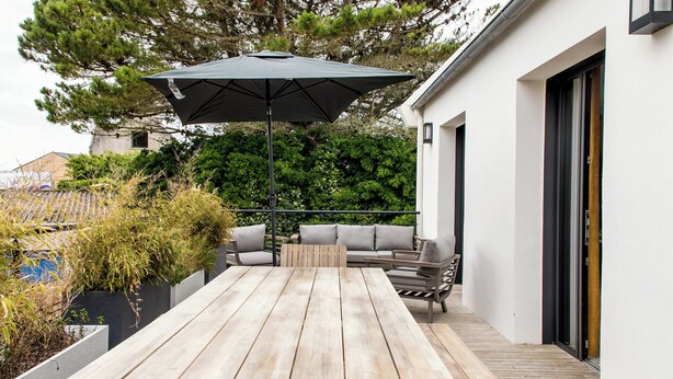 A wooden table with an umbrella, outdoor seating, and a white building in the background. - La Maison des Dunes - Plage à Pied (Cléder)