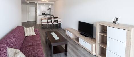 A modern living room with a sofa, a coffee table, a TV, and a kitchen area in the background.