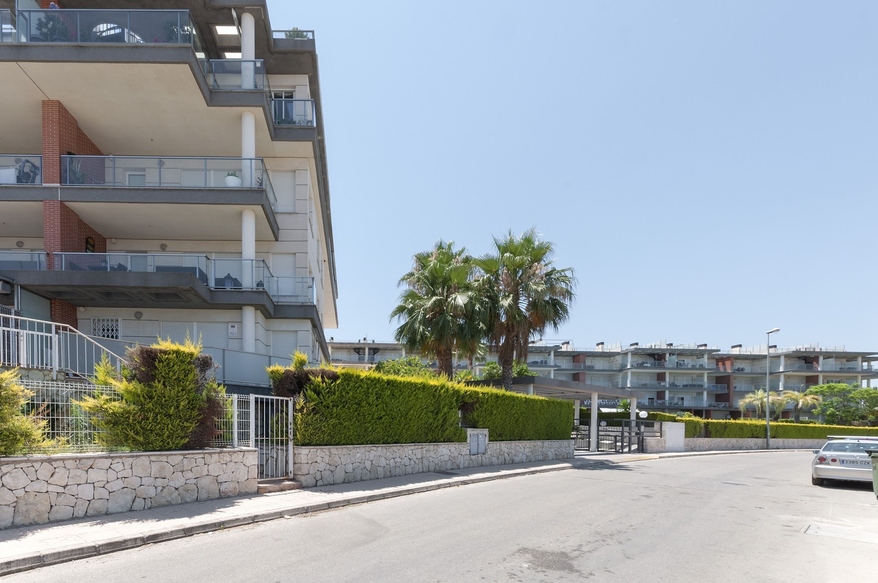 A multi-story residential building with balconies, a stone retaining wall, and palm trees.